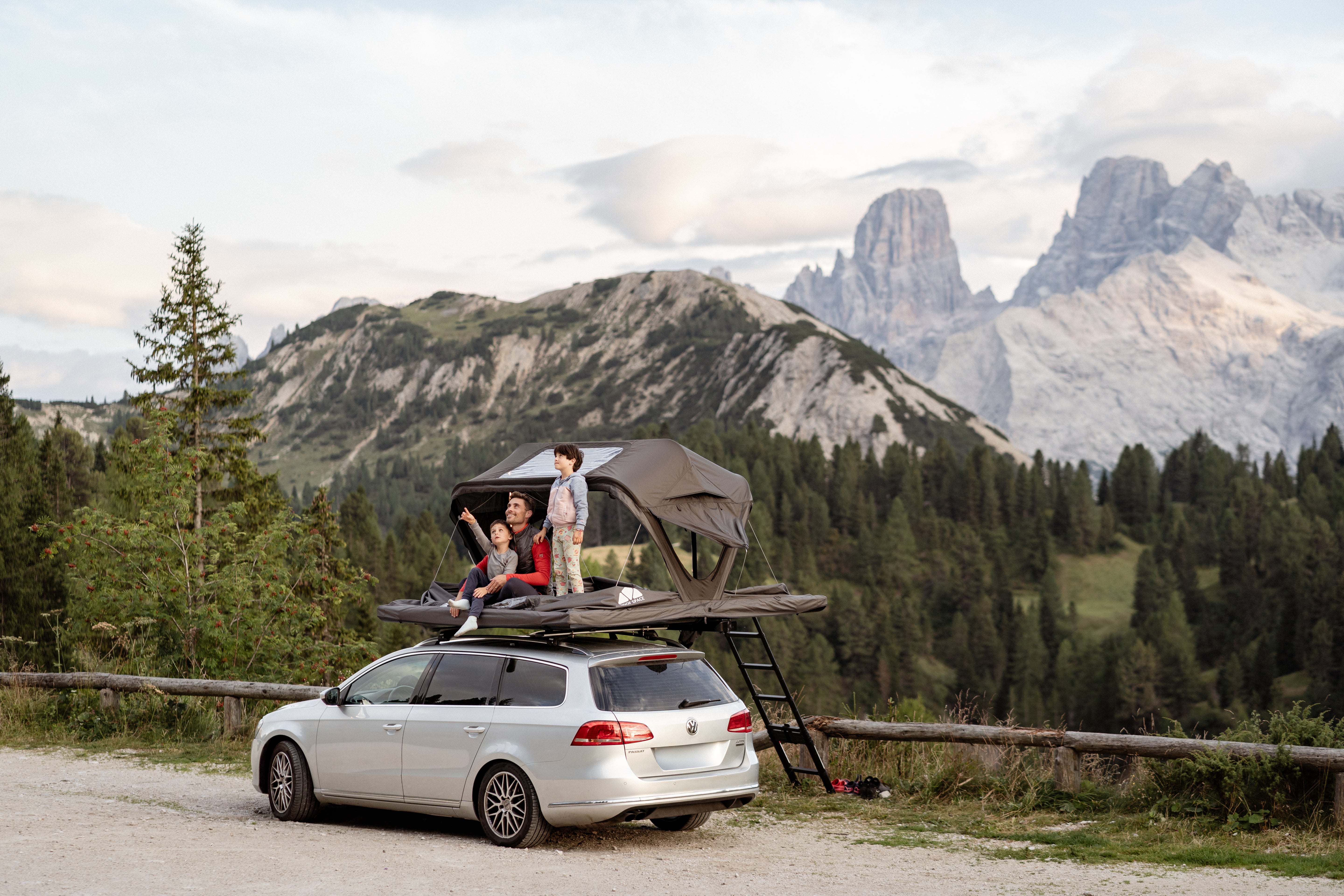 Familie mit Zelt auf silbernem VW Passat vor Bergkulisse. Vater und Kinder sitzen/stehen im Zelt und schauen nach links. Ruhige, helle Atmosphäre.
