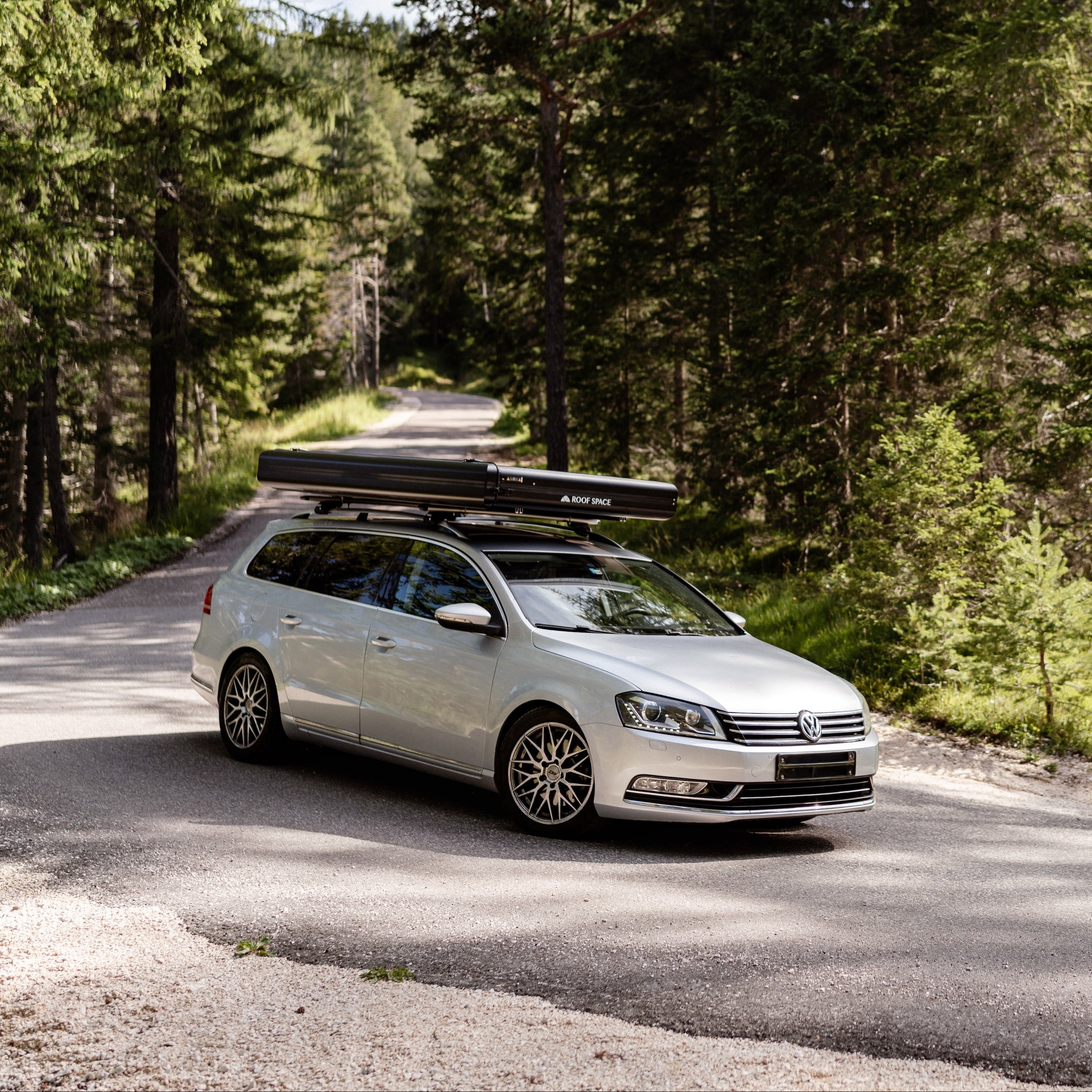 Silberner VW Passat mit Dachzelt auf gewundener Straße durch einen hellen, sonnigen Wald. Auf dem Zelt steht "ROOF SPACE".