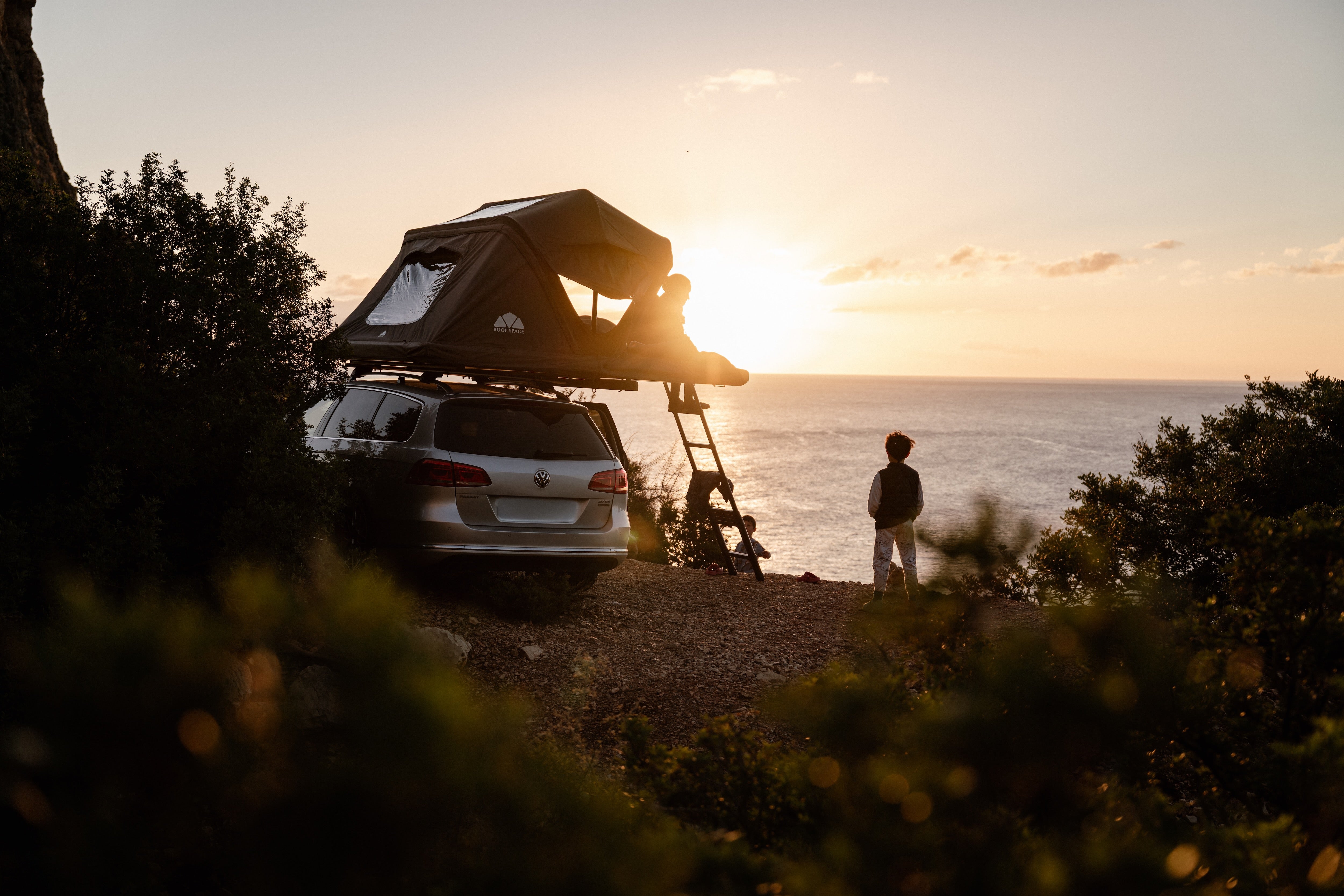 Silberner VW Passat mit Dachzelt am Hang, Kinder auf Leiter und am Ufer, Sonnenuntergang über dem Meer im Hintergrund. "ROOF SPACE" auf dem Zelt sichtbar.