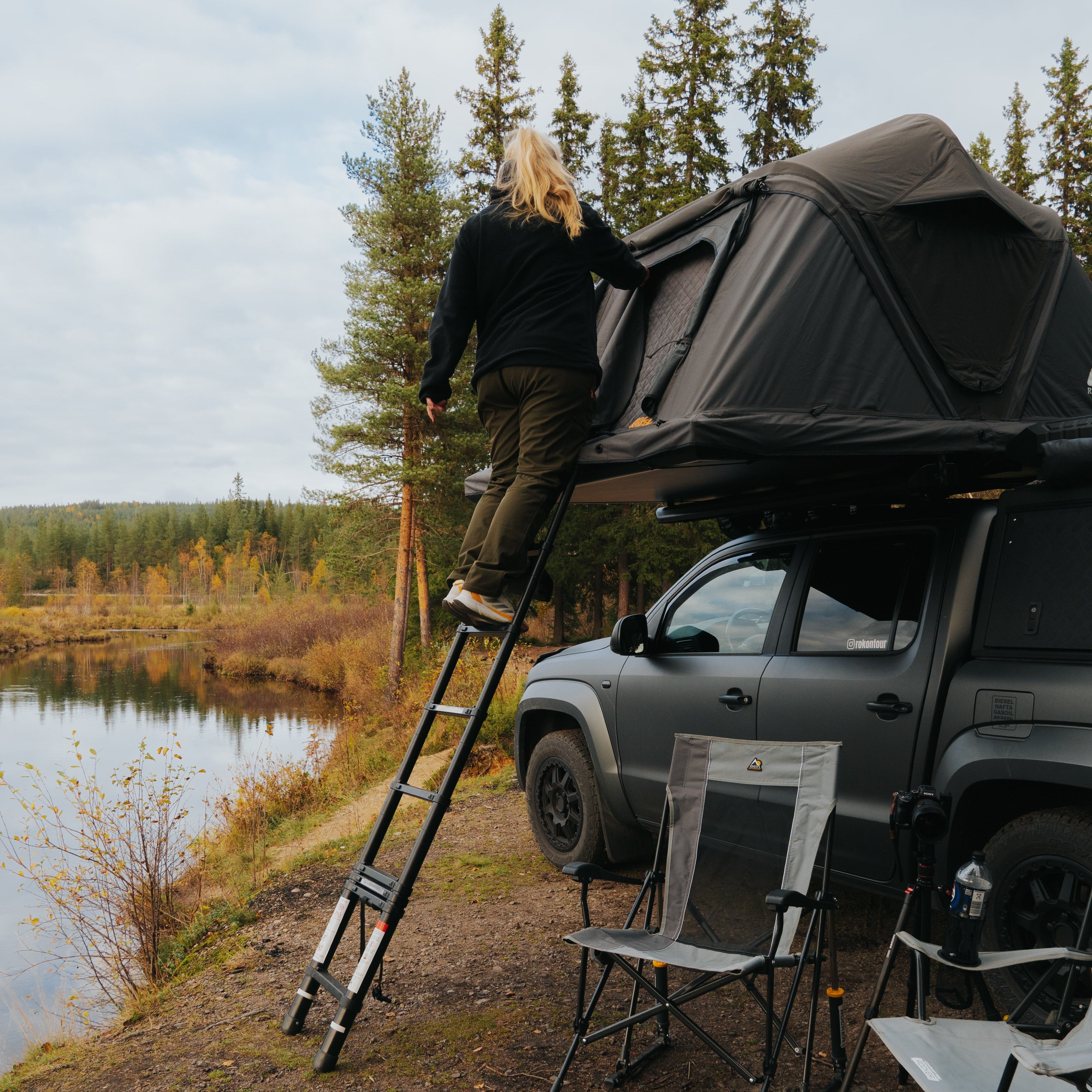 Eine blonde Frau steigt über eine Leiter auf ein graues Auto mit Dachzelt am Ufer eines Sees. Im Hintergrund sind Bäume.