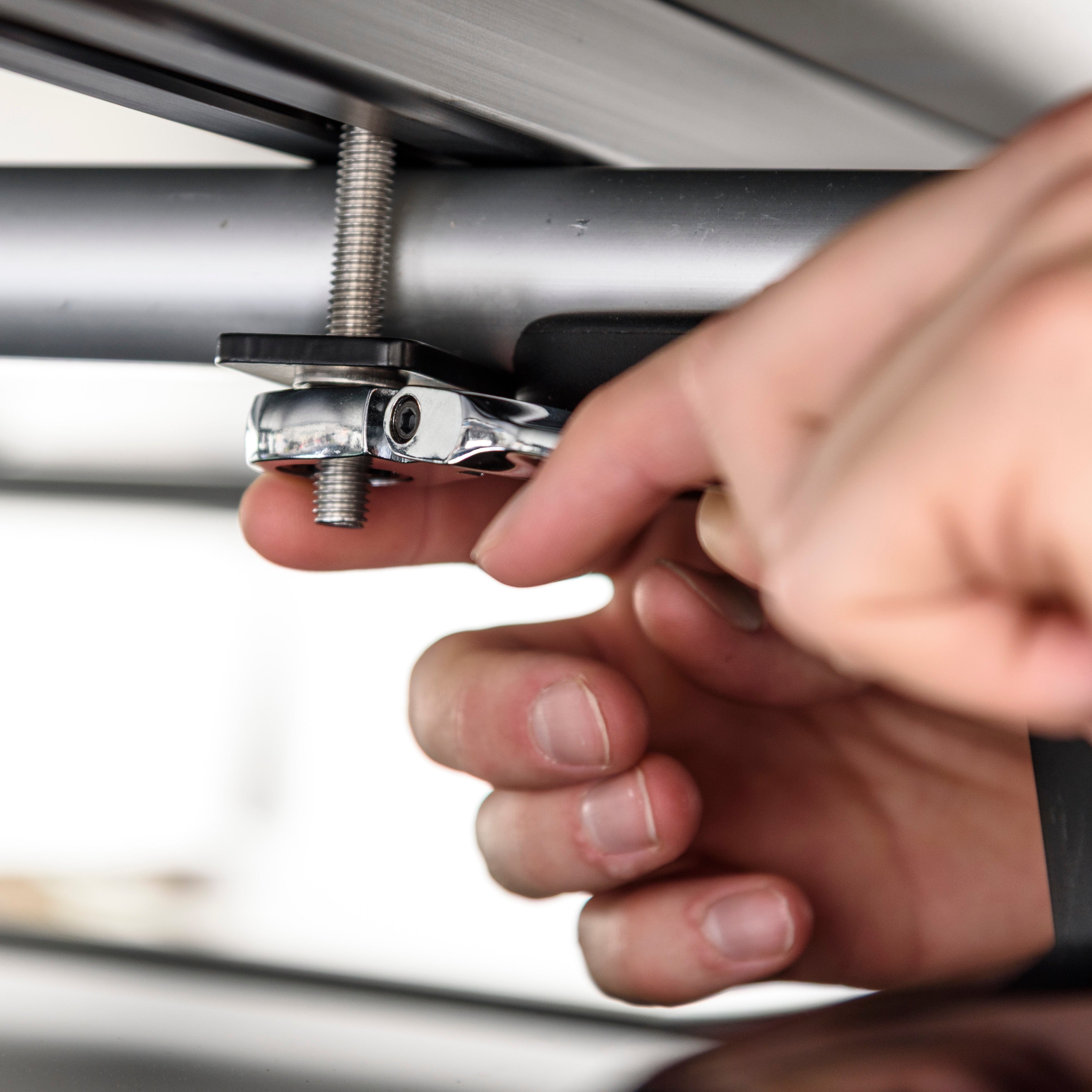 Nahaufnahme: Hand bedient einen verchromten Spannhebel an einer Gewindestange unterhalb einer Metallstruktur, helle, neutrale Farbtöne.