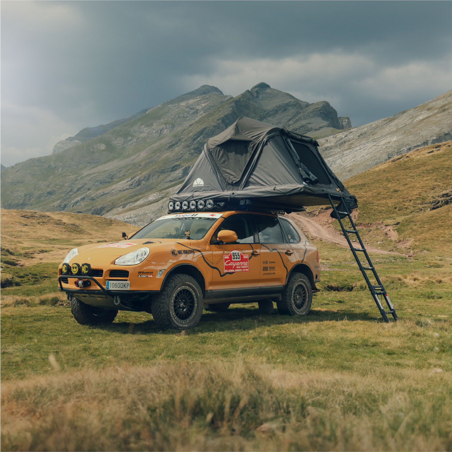 Geländegängiger, orangefarbener Porsche Cayenne mit Dachzelt vor einer Berglandschaft, Leiter angelehnt. Aufschrift "Cayenne Epic Trophy 955". Bewölkter Himmel, gedämpftes Licht.