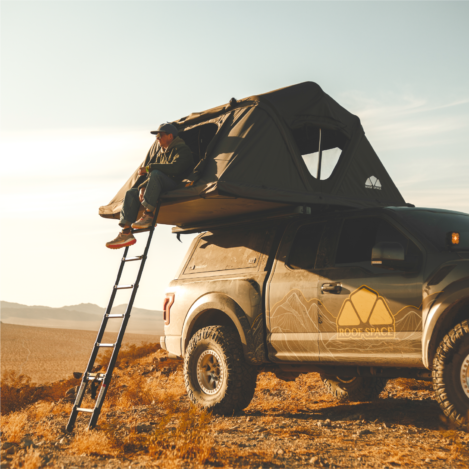 Ein Mann sitzt auf einem Dachzelt auf einem Pickup-Truck in der Wüste. Eine Leiter steht links. Das Logo „ROOF SPACE“ ist sichtbar.