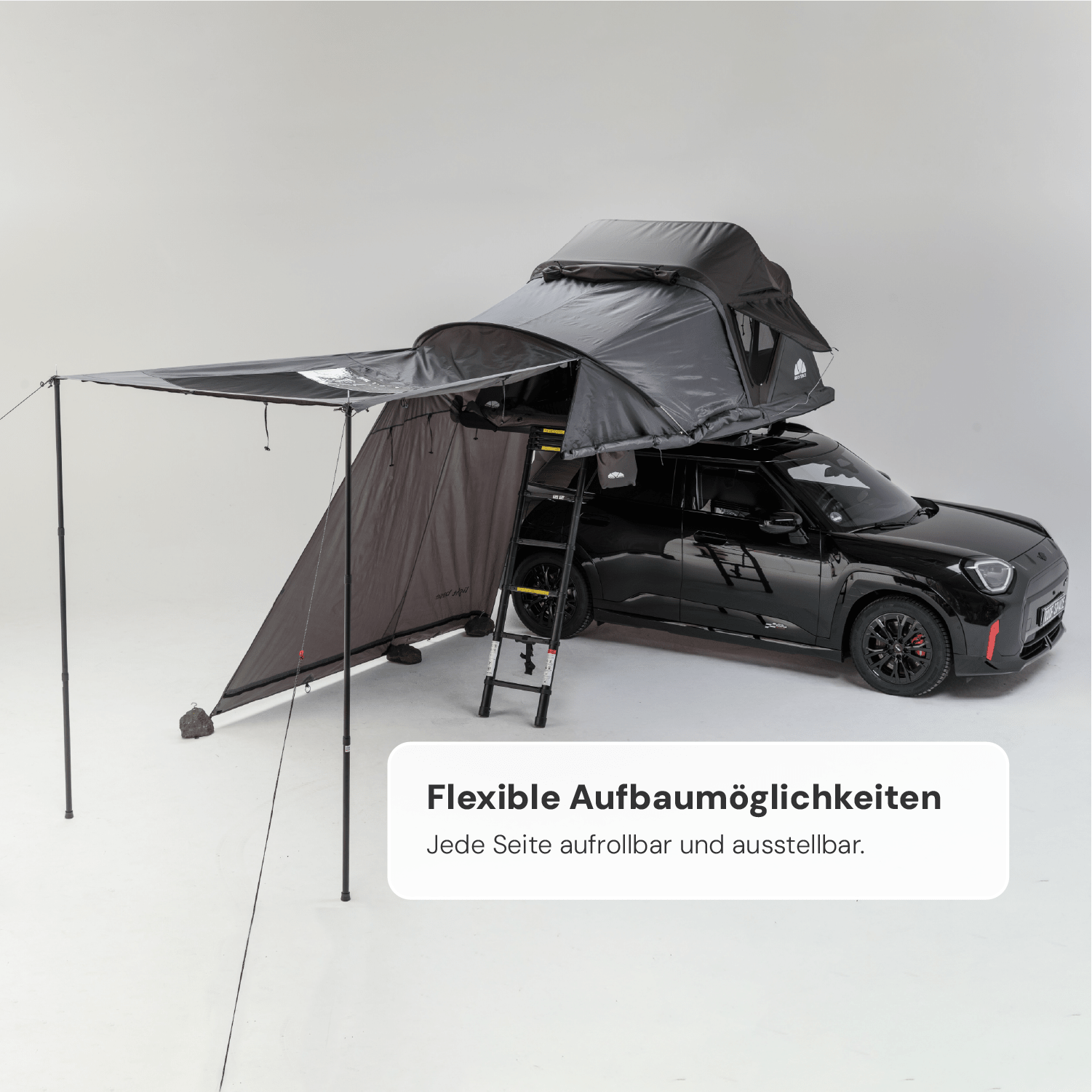 Schwarzer Mini Cooper mit grauem Dachzelt und Markise in einem hellen Studio. Text: "Flexible Aufbaumöglichkeiten. Jede Seite aufrollbar und ausstellbar."