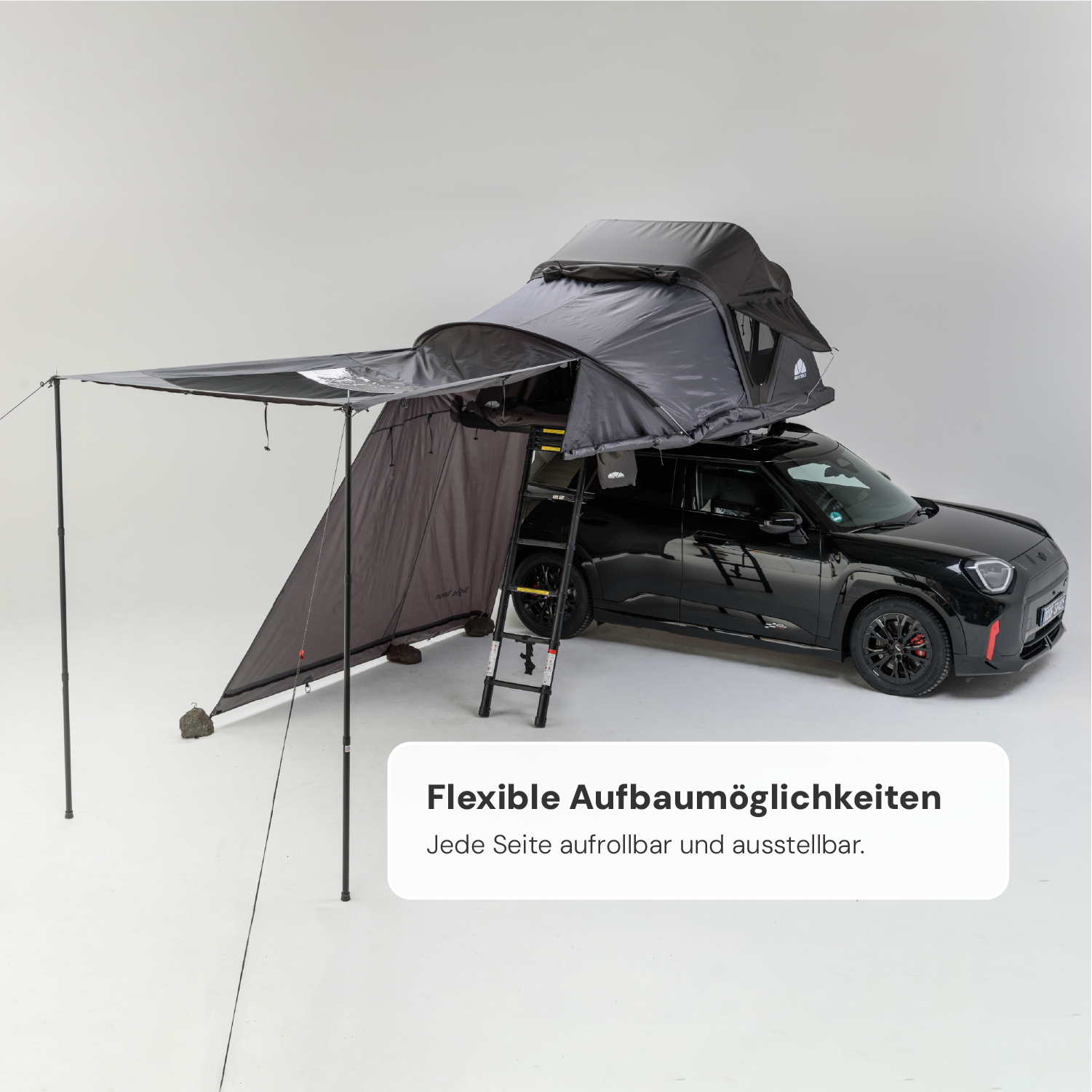 Schwarzer Mini Cooper mit grauem Dachzelt und Markise in einem hellen Studio. Text: "Flexible Aufbaumöglichkeiten. Jede Seite aufrollbar und ausstellbar."