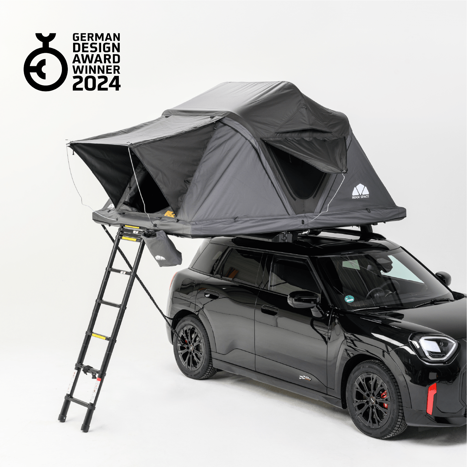 Schwarzer Mini Countryman mit Dachzelt und Leiter auf weißem Hintergrund. Das Zelt ist grau und trägt das "German Design Award Winner 2024"-Logo.