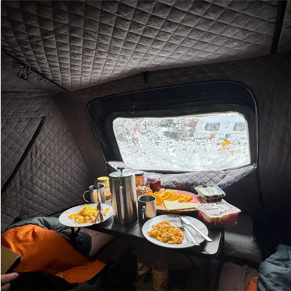 Cozy breakfast in the tent: dishes with scrambled eggs, sausage, cheese, and fruit on a small table set against a snowy window view.