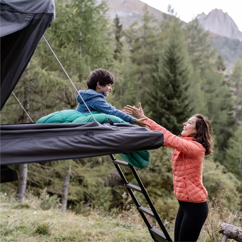 A child sitting at the open panoramic mode of the rooftop tent Roof Space 4 while the mother stands by the ladder below reaching out her arms in a scenic natural setting.