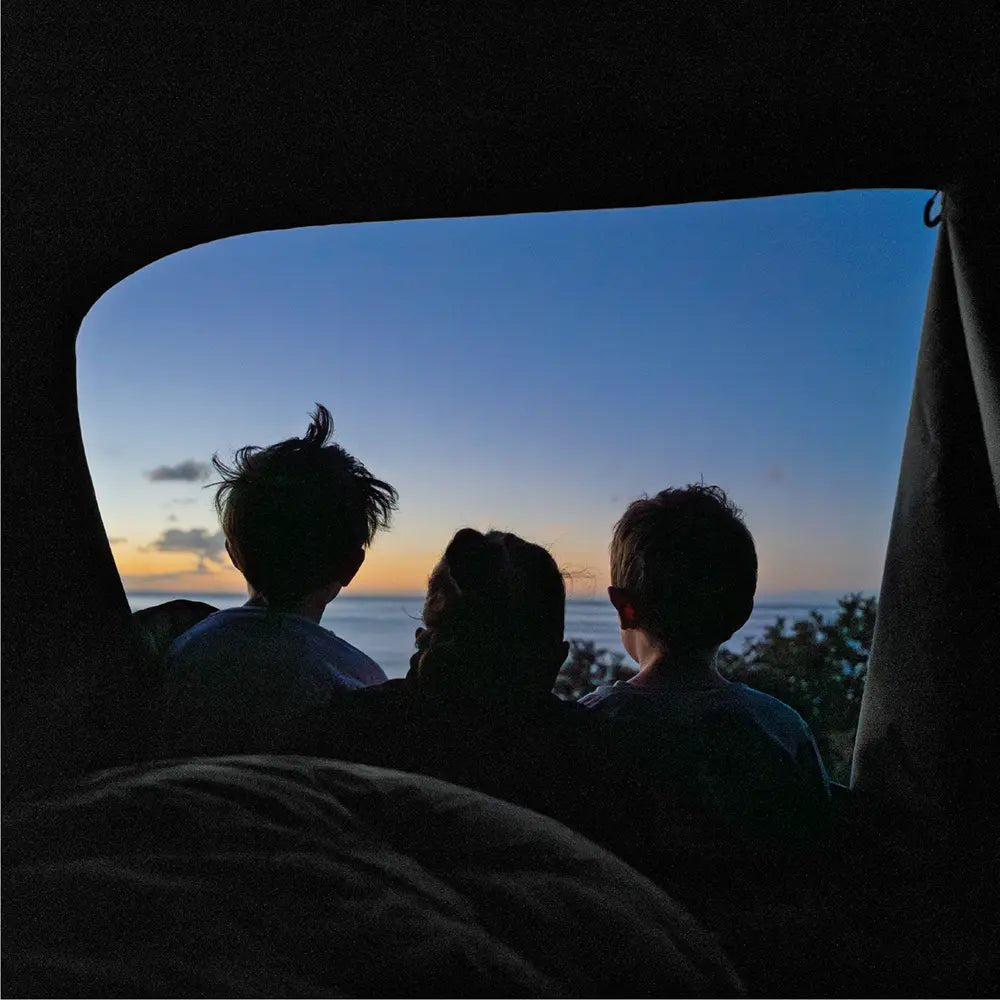A mother and two children laying inside the open rooftop tent Roof Space 4, watching the dusk sky over the ocean.