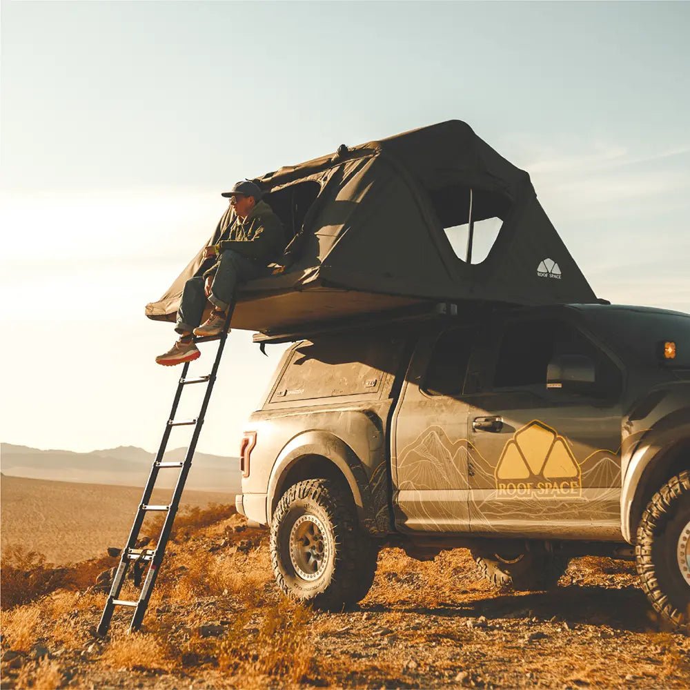 A man relaxing inside a Roof Space rooftop tent mounted on an off-road vehicle with the yellow logo in a barren landscape.