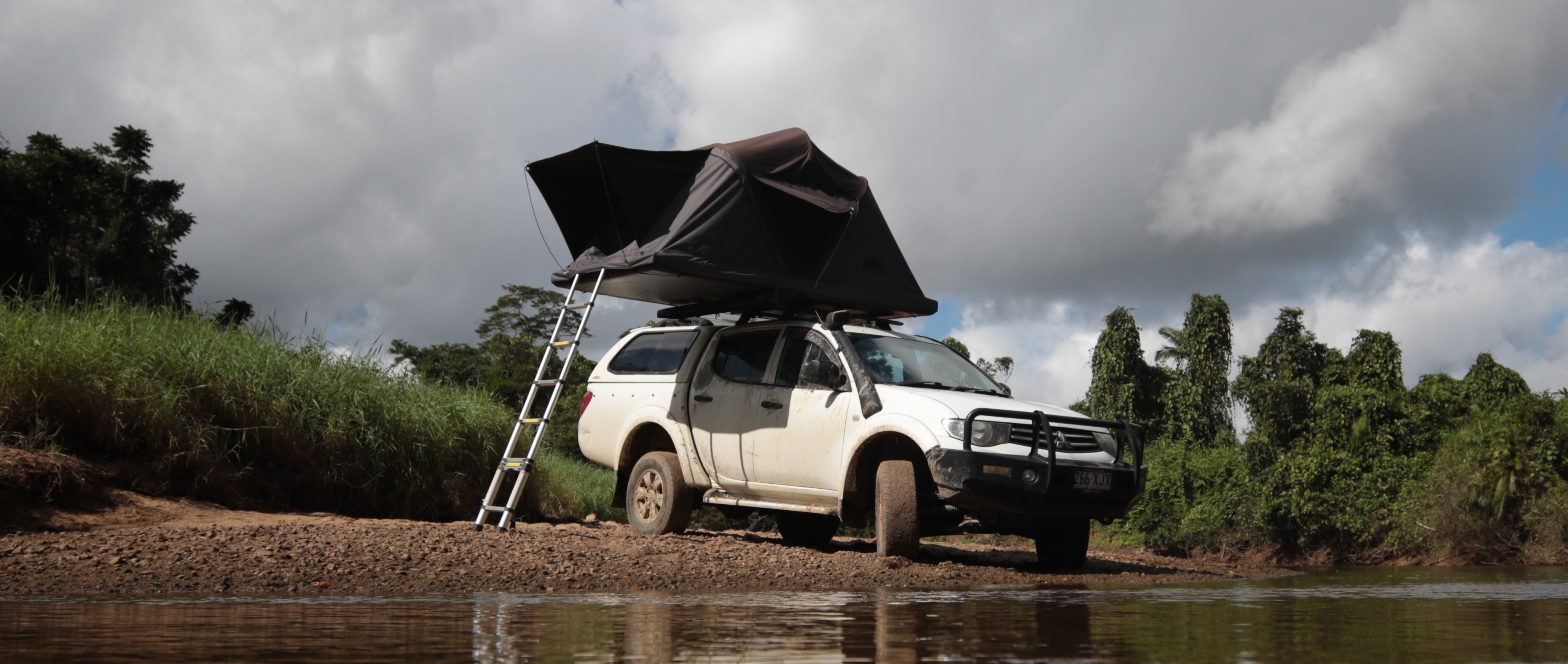 Ein weißer Geländewagen mit Dachzelt steht am Ufer eines Flusses unter bewölktem Himmel. Eine Leiter führt zum Zelt. Nummernschild sichtbar: 266 XJX.