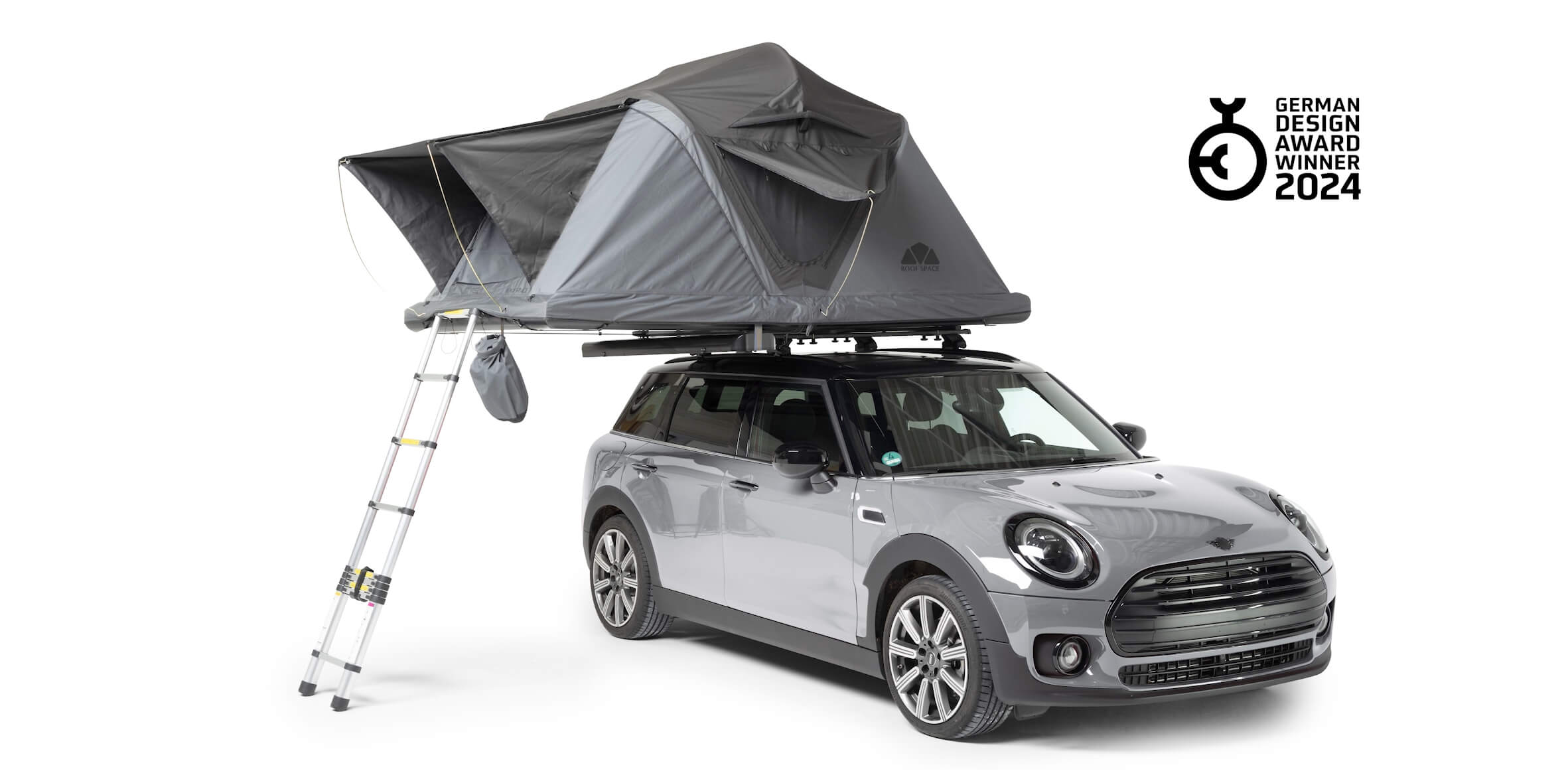 Ein grauer Mini Clubman mit einem grauen Dachzelt und einer Leiter vor weißem Hintergrund. Rechts oben steht "German Design Award Winner 2024".