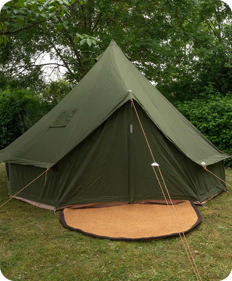 Ein olivgrünes Tipi-Zelt steht auf einer Wiese vor Bäumen. Braune Seile spannen das Zelt. Ein brauner Teppich liegt vor dem Eingang. Ruhige, natürliche Atmosphäre.