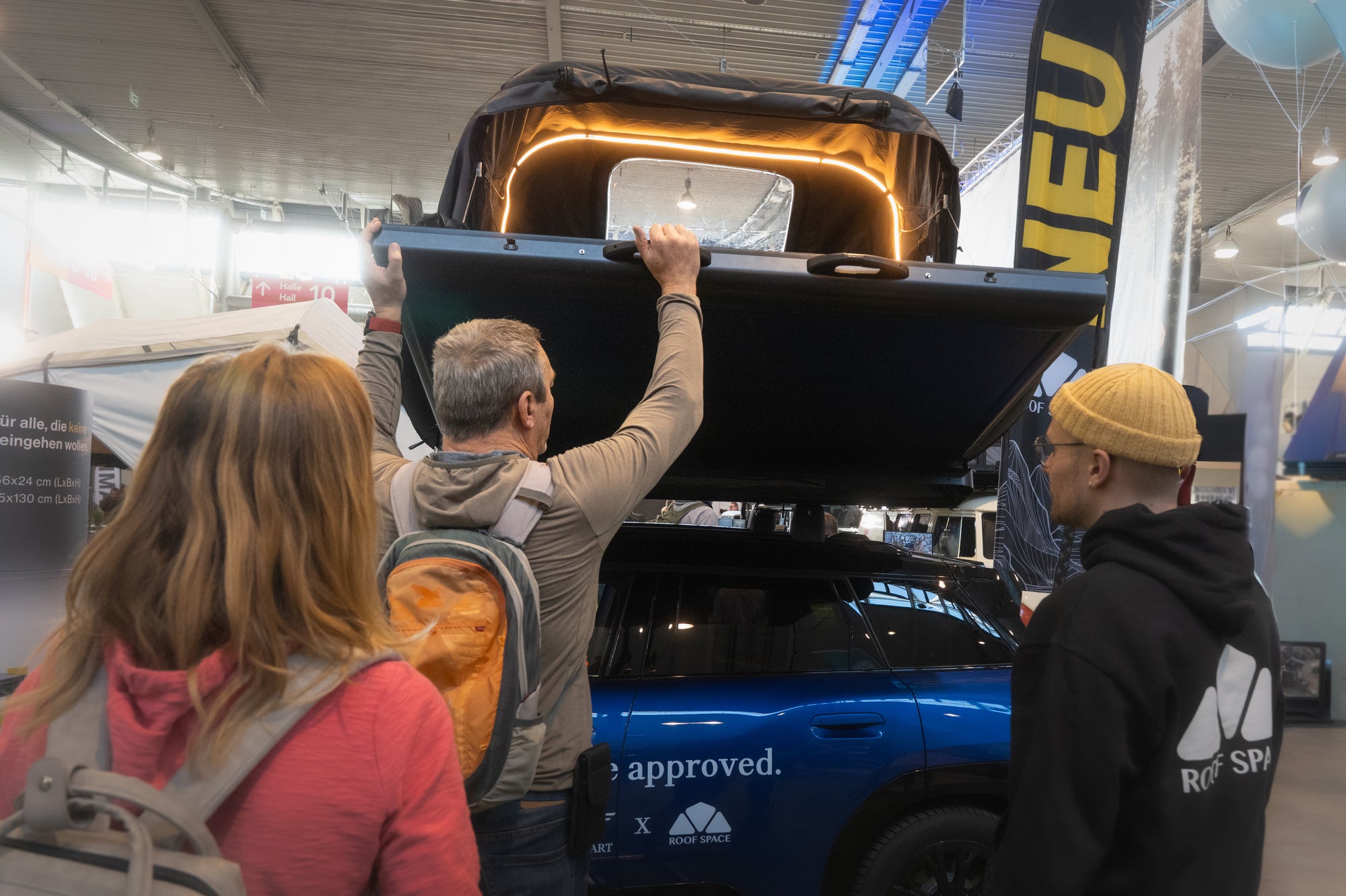 Auf einer Messe öffnet ein Mann ein Dachzelt auf einem blauen Auto; im Vordergrund stehen zwei weitere Personen. Im Hintergrund sieht man ein Banner mit der Aufschrift "NEU".