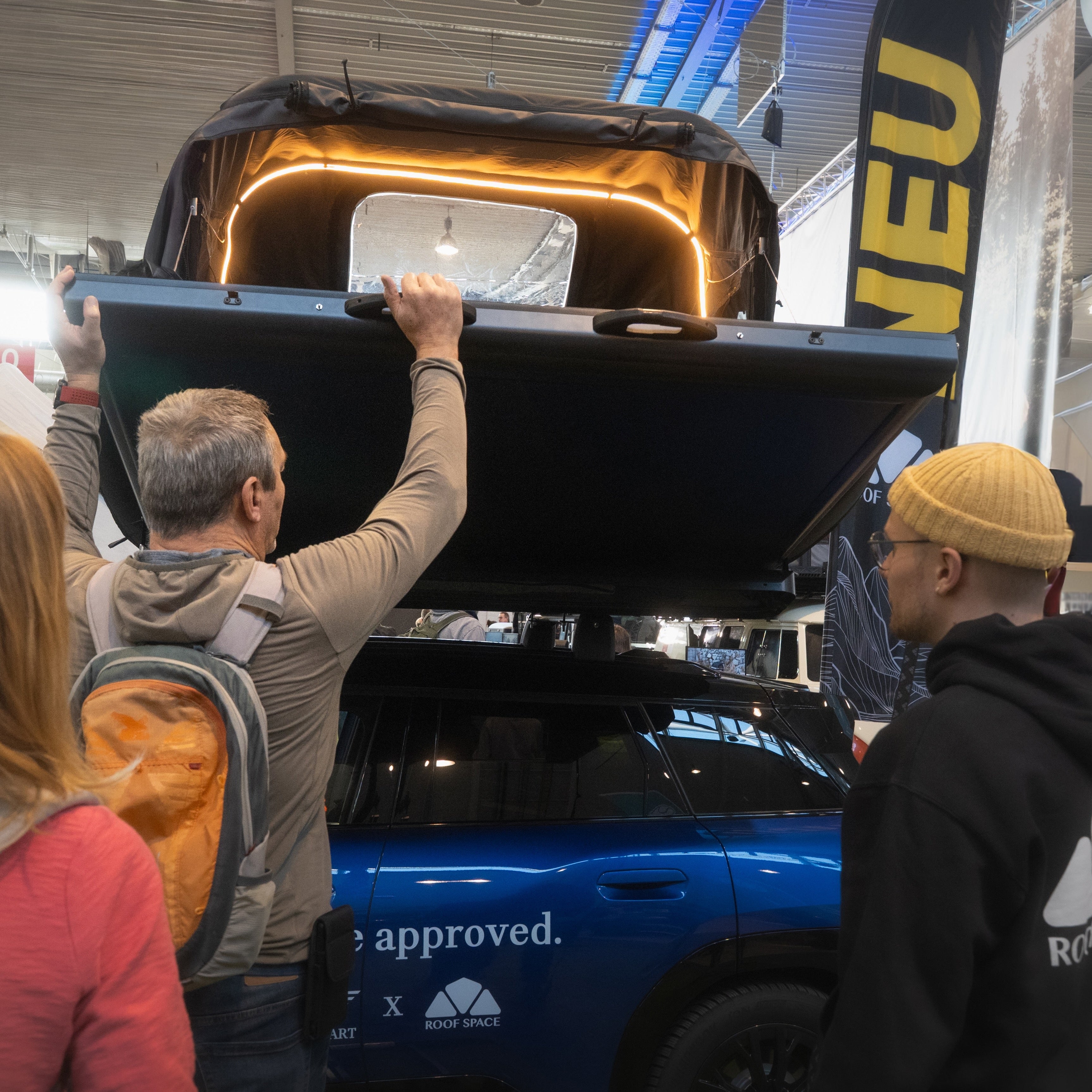 Eine dunkelblaue Auto mit Dachzelt, das von einem Mann geöffnet wird. Ein junger Mann mit gelber Mütze schaut zu. Im Hintergrund Banner mit der Aufschrift "NEU".