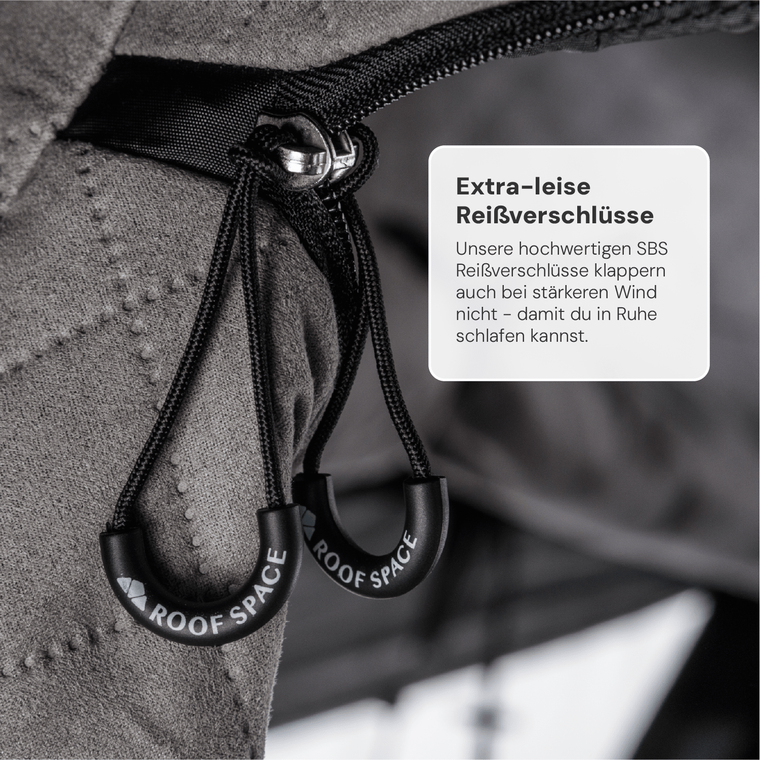 Nahaufnahme: Zwei schwarze Reißverschlussanhänger mit "ROOF SPACE" Schriftzug hängen an einem grauen Stoff. Textfeld rechts: "Extra-leise Reißverschlüsse".