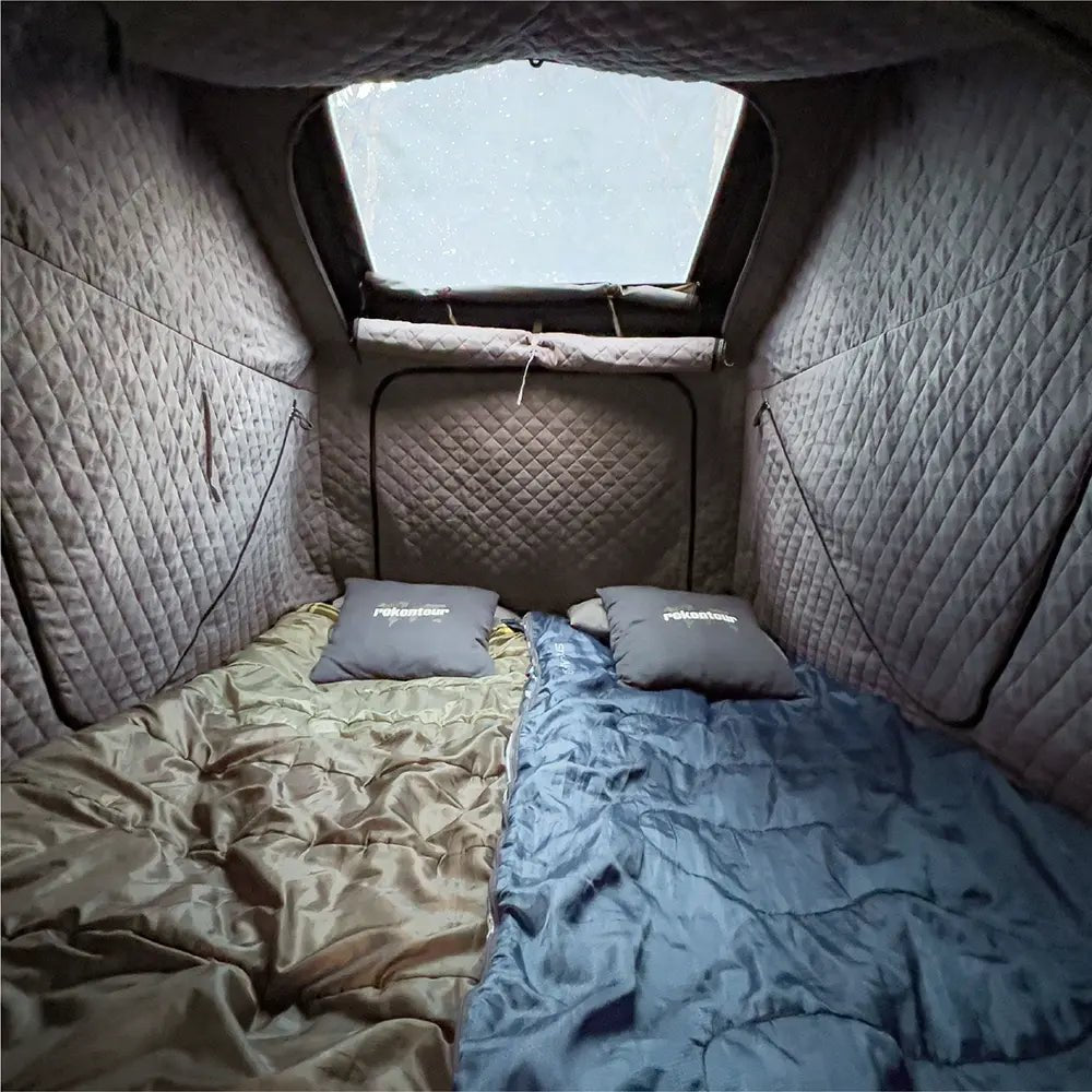 Interior view of a tent with two beds—one beige and one blue-each featuring a "rokontour" pillow. The tent walls are quilted.
