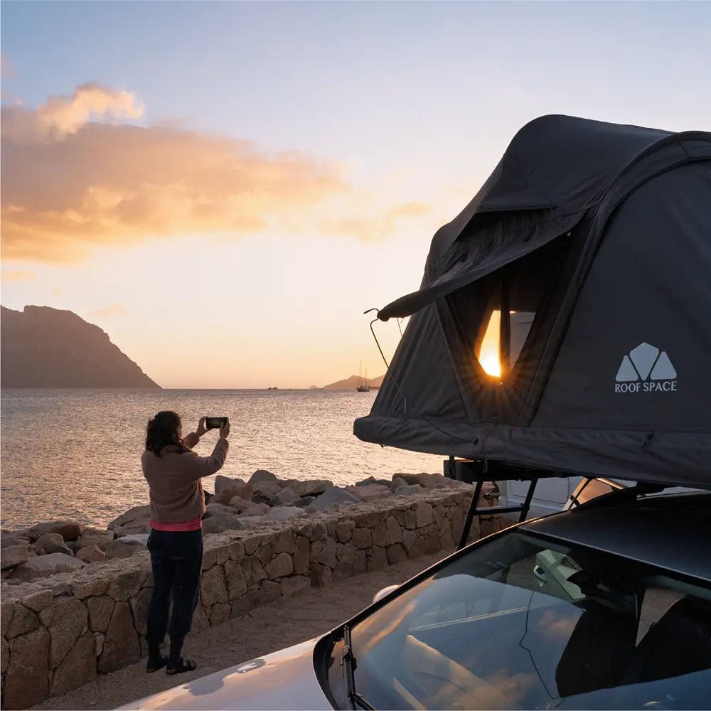A person using a smartphone to capture the ocean sunset while standing next to their rooftop tent setup "Roof Space 4" on the beach.