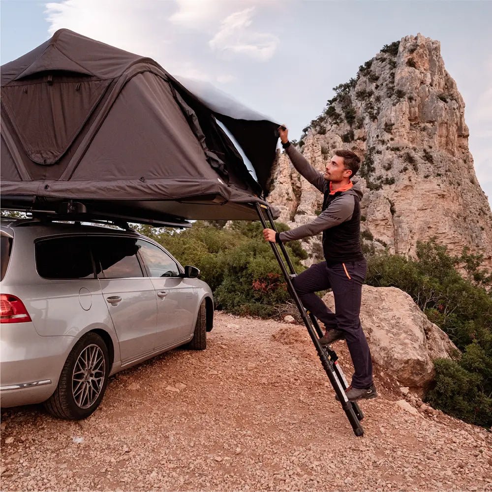 A man climbing the ladder to set up the porch awning on the Roof Space 4 rooftop tent mounted on a VW Passat along a mountainous coastline.