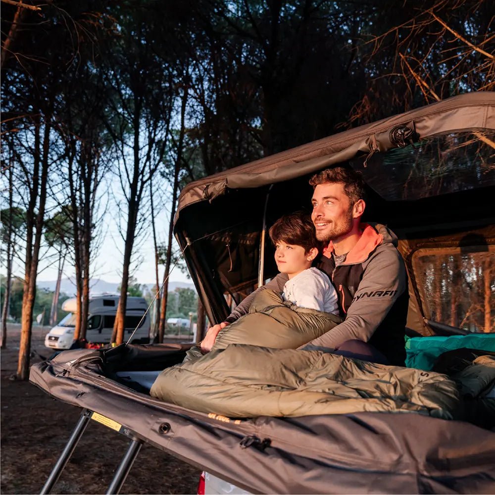 A father and child cozy in a sleeping bag inside the open rooftop tent Roof Space 4, enjoying the sunset together.