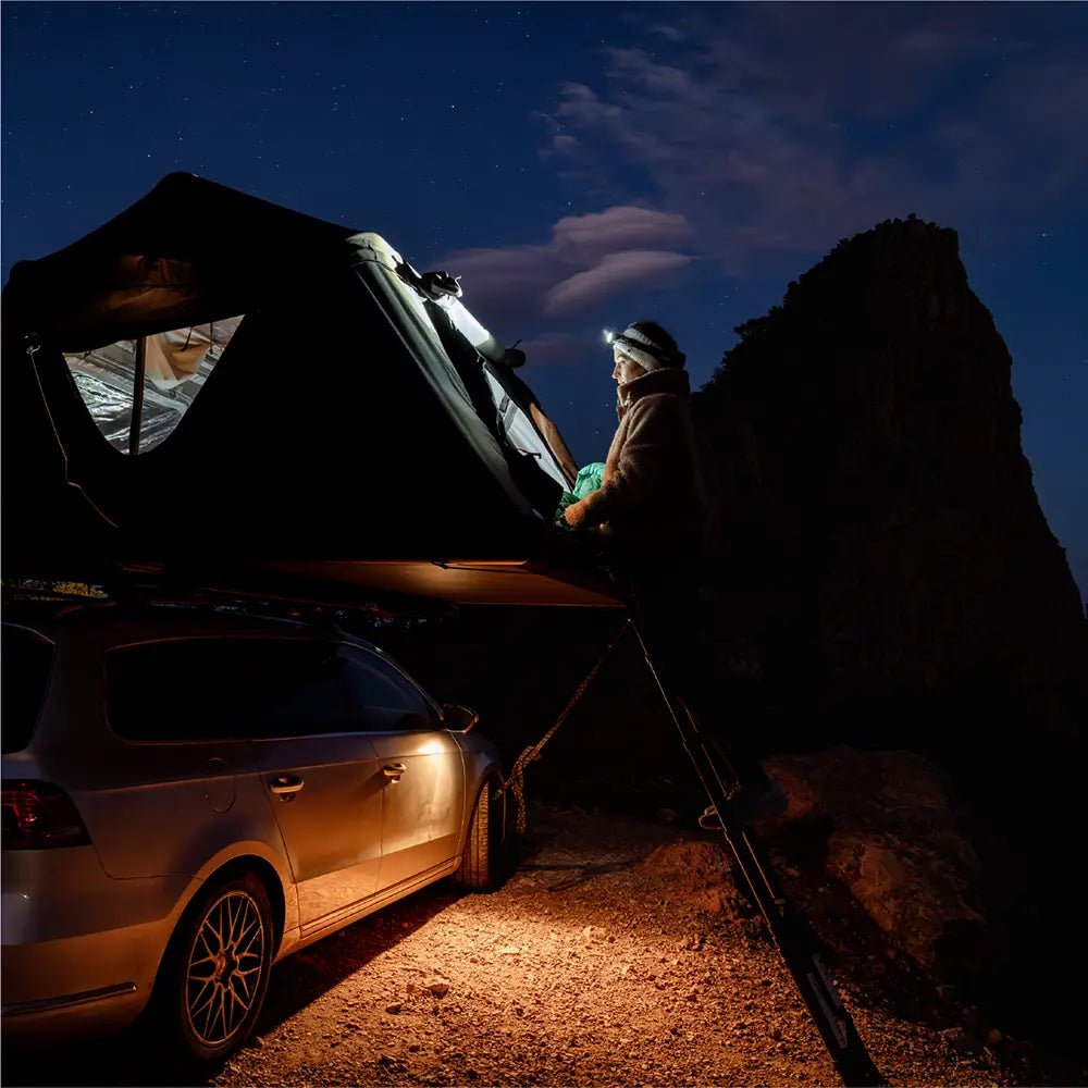 A person standing on the ladder with a flashlight at night, about to climb into the illuminated rooftop tent.