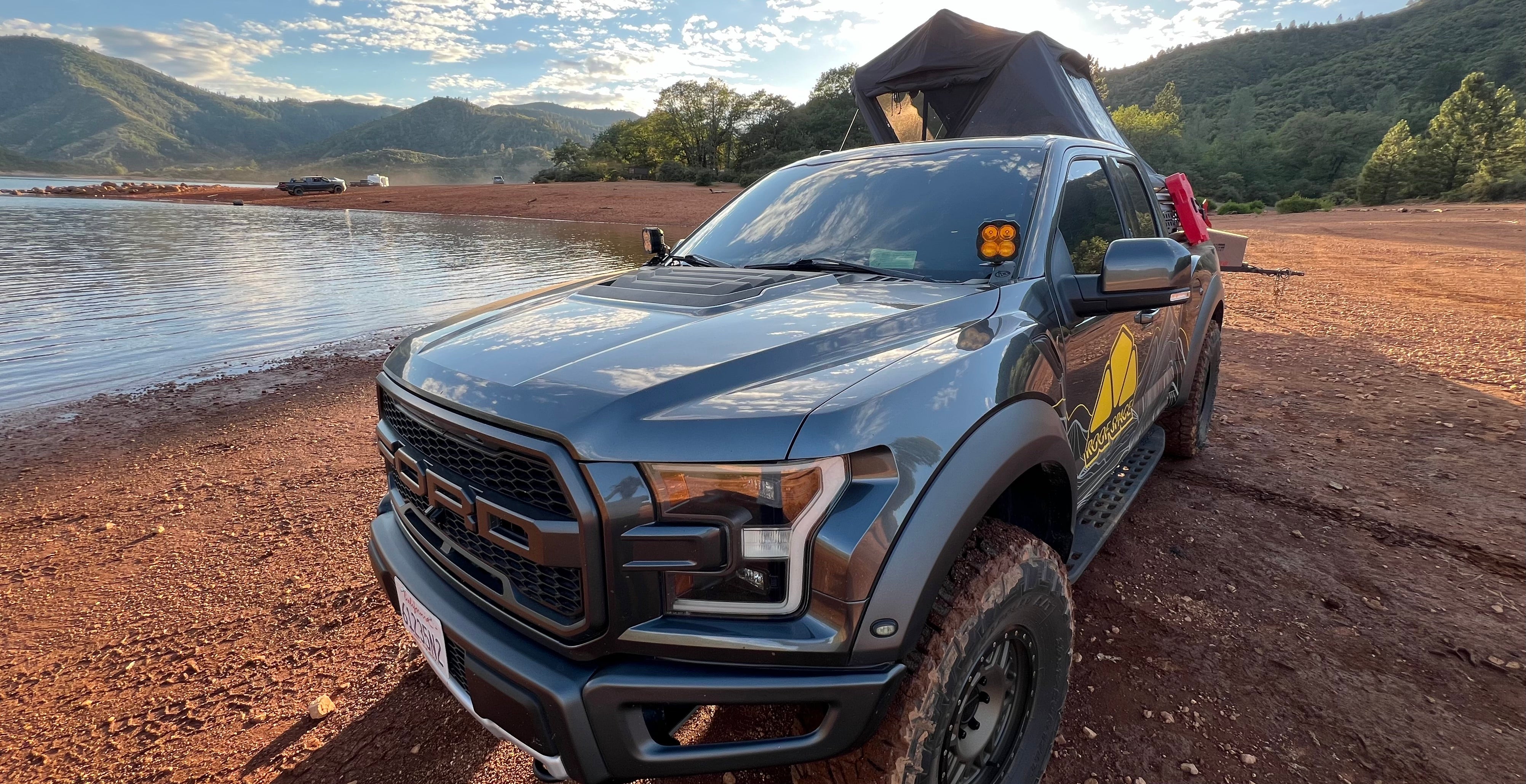 Ein grauer Ford Raptor mit Dachzelt steht am Ufer eines Sees. Berge im Hintergrund, sonnendurchflutete Szene. Schriftzug "Rock Stack" auf der Fahrzeugseite sichtbar.