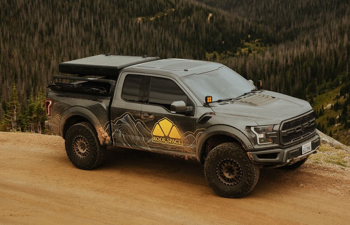 Grauer Ford Raptor Pickup-Truck mit Dachzelt auf schmutziger Straße vor bewaldetem Berghang. "ROOF SPACE" Logo auf der Seite.