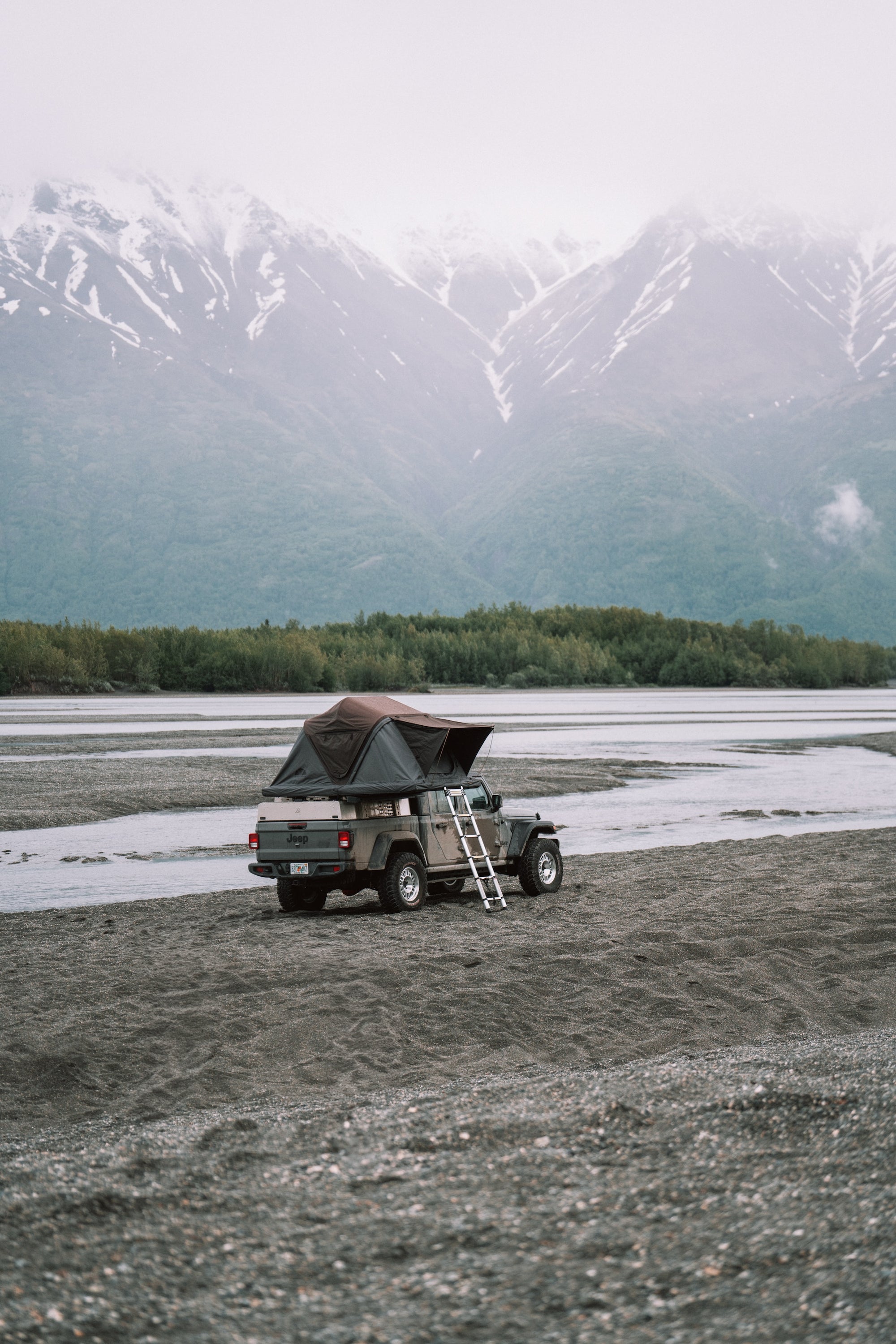 Ein grauer Jeep Gladiator mit Dachzelt steht auf einer Kiesfläche; im Hintergrund sind Berge mit Schnee zu sehen. Trübe Atmosphäre.