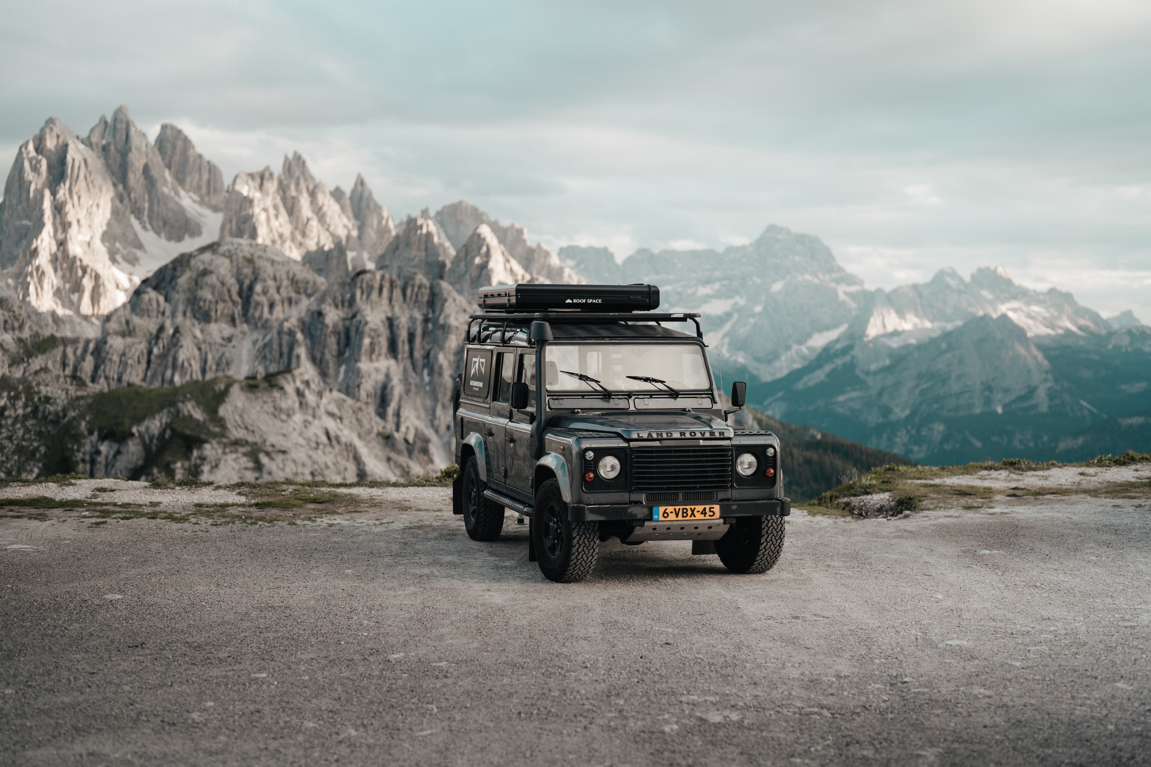 Ein schwarzer Land Rover Defender parkt vor einer Bergkulisse unter bewölktem Himmel. "ROOF SPACE" ist auf dem Dachzelt zu lesen, das Kennzeichen ist "6-VBX-45".