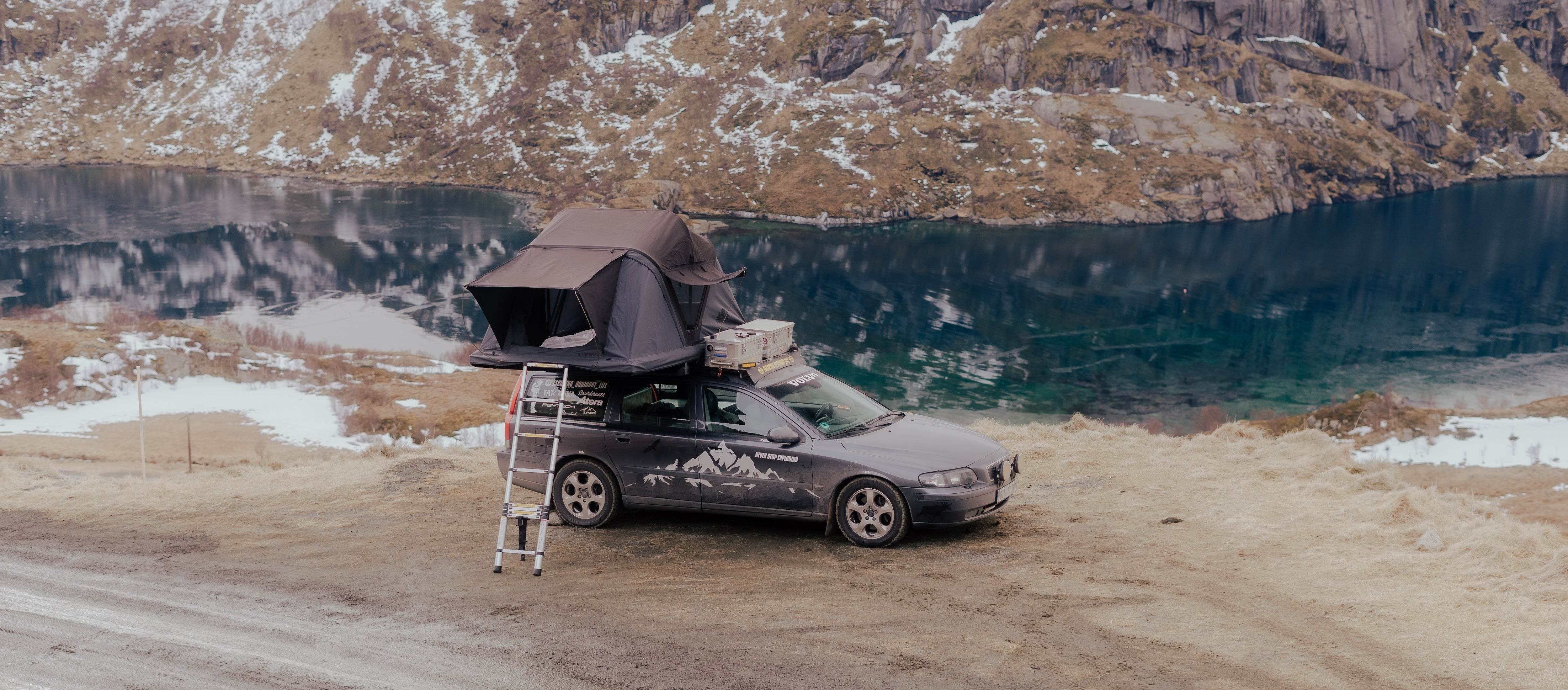 Ein grauer Volvo mit Dachzelt und Leiter steht am Ufer eines Sees vor bergiger Landschaft. Auf der Seite des Autos steht "Never Stop Exploring".