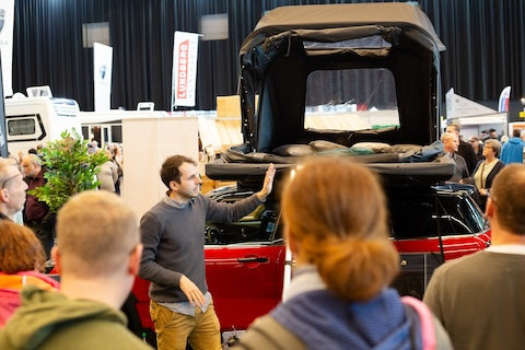 Auf einer Messe erklärt ein Mann vor einem roten Auto mit Dachzelt Details vor interessiertem Publikum. "LUNDBERG" ist im Hintergrund auf einem Schild zu lesen.