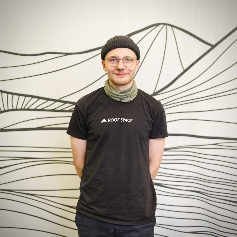 Ein Mann mit Brille, Mütze und Halswärmer steht vor einer Wand mit Bergmotiv. Er trägt ein schwarzes T-Shirt mit dem Text "ROOF SPACE".