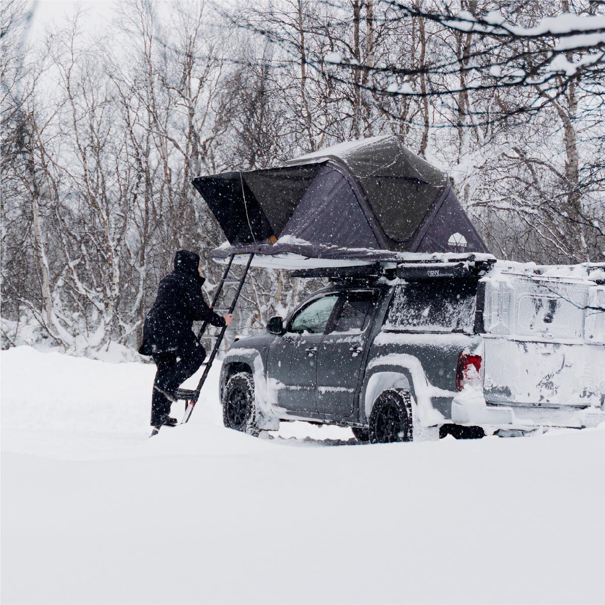 Winterlandschaft mit schneebedecktem grauen Pickup-Truck; Person steigt Leiter zum Zelt auf dem Dach hoch. Schneefall, Bäume im Hintergrund.
