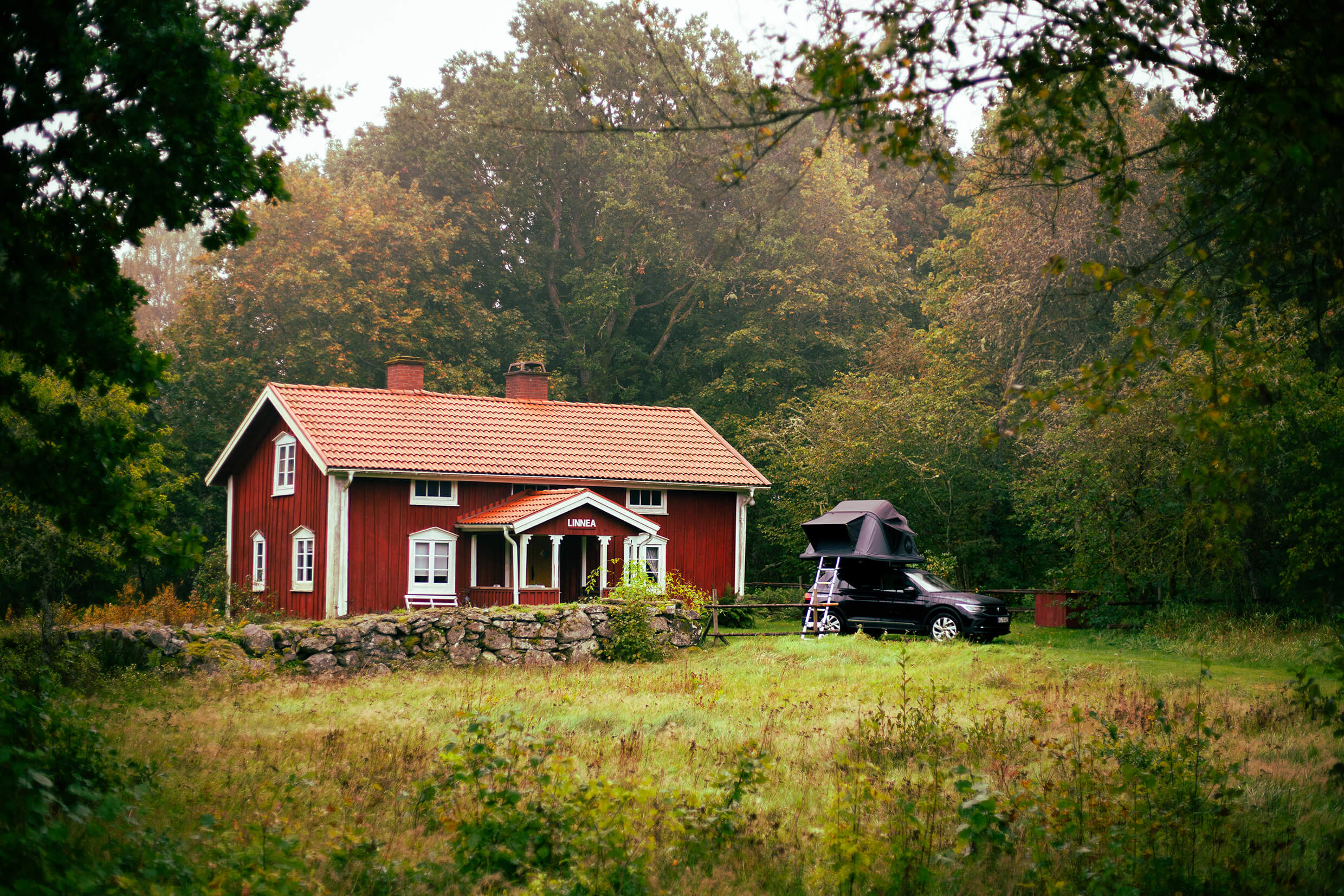 Rotes Holzhaus mit weißen Fensterrahmen im Grünen; ein schwarzes Auto mit Dachzelt steht rechts davon. "LINNEA" steht über dem Eingang.