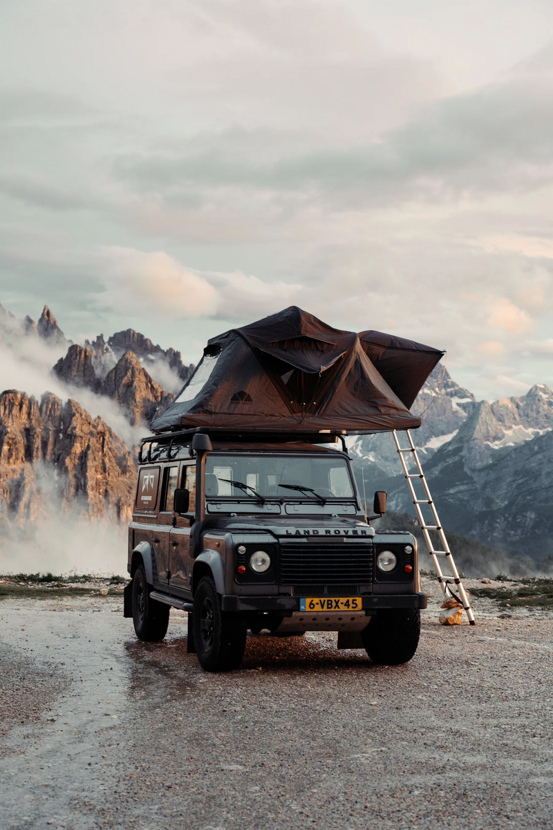 Ein grauer Land Rover Defender mit Dachzelt vor einer Bergkulisse im bewölkten Morgenlicht. Das Kennzeichen "6-VBX-45" ist sichtbar.