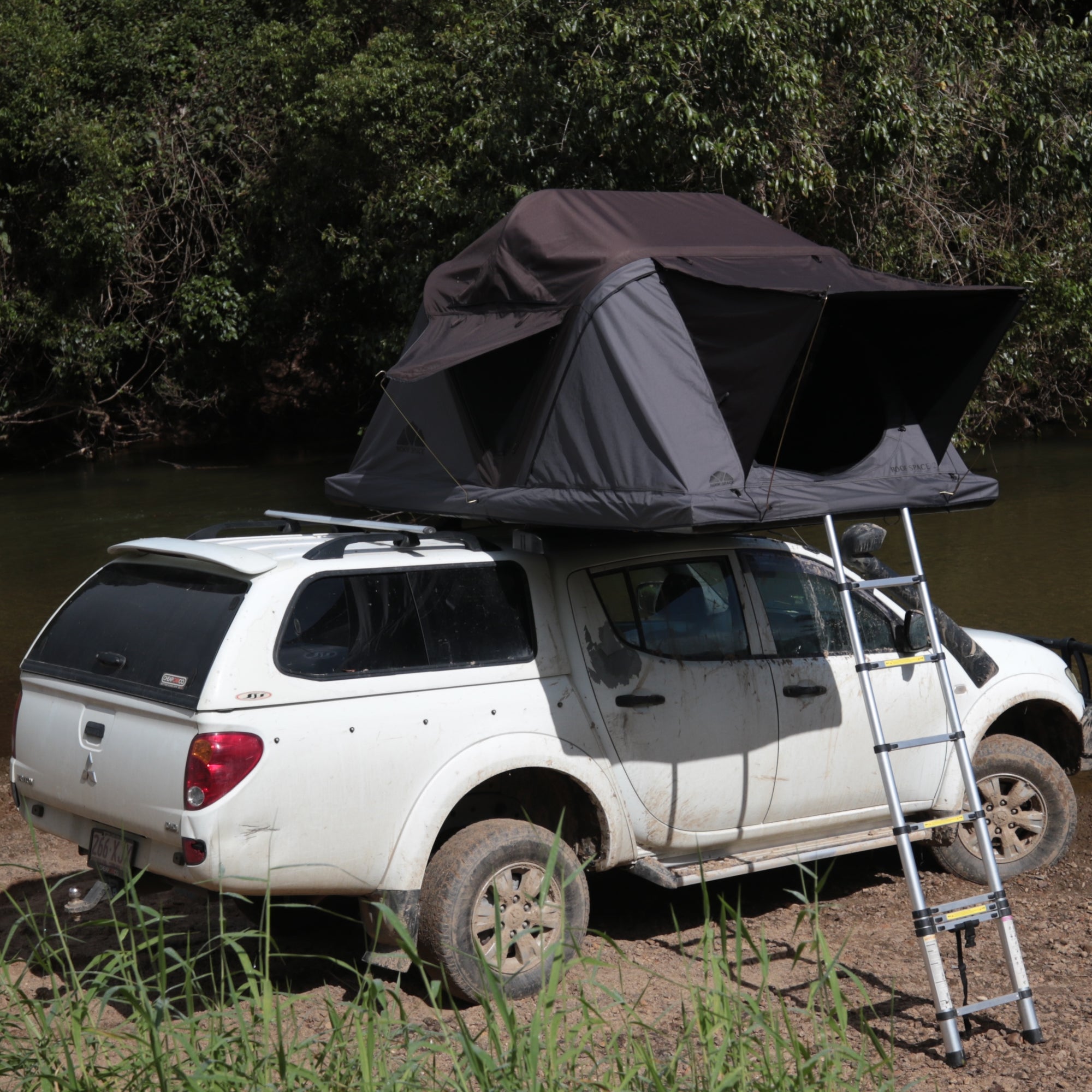 Ein weißer, schmutziger Pick-up mit Dachzelt steht am Ufer, eine Leiter lehnt am Wagen. Im Hintergrund Bäume und Wasser. "266-XJJ" ist auf dem Nummernschild lesbar.