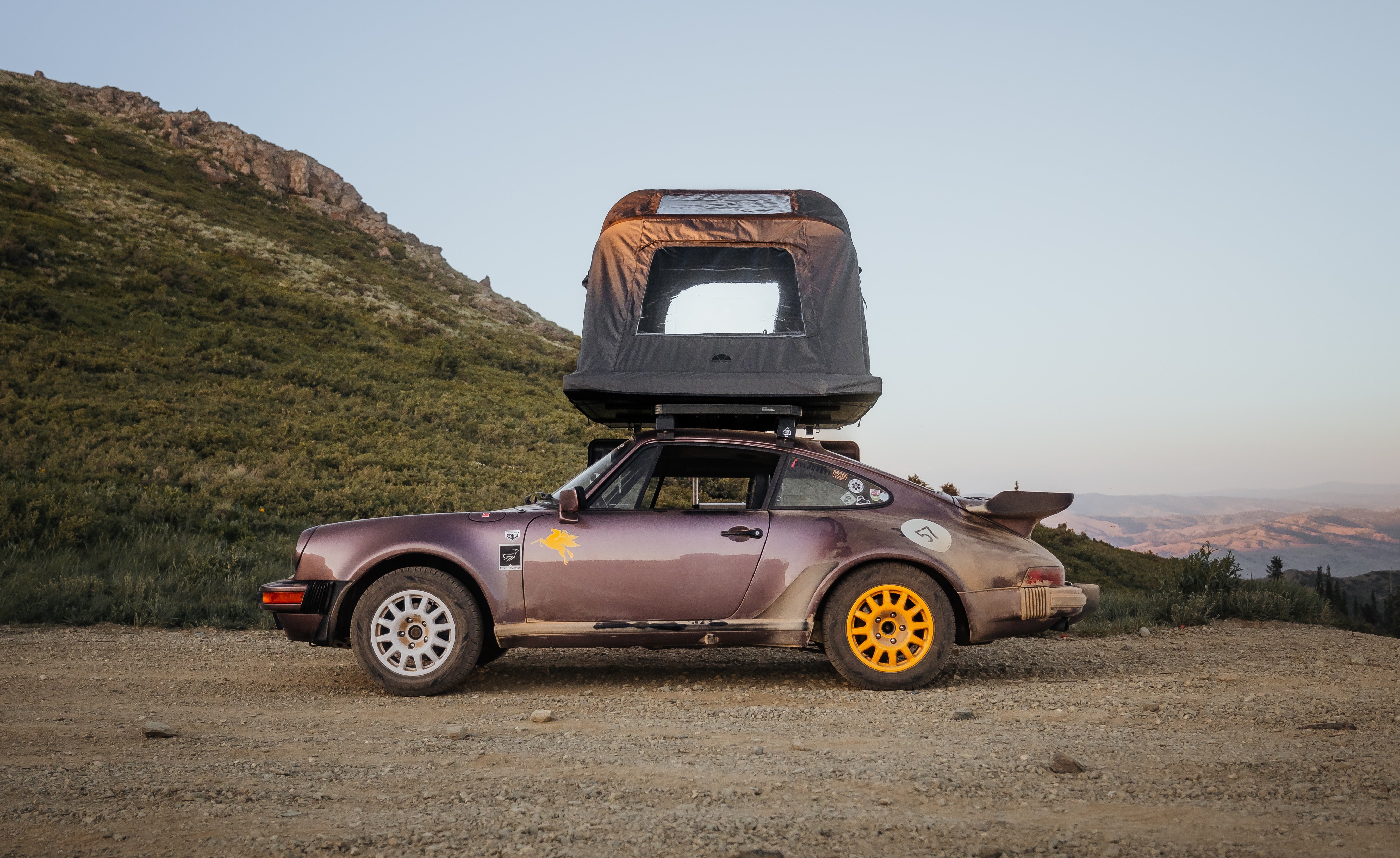 Ein auberginefarbener Porsche 911 mit Dachzelt steht auf einem Schotterweg, im Hintergrund Berge und Hügel. Gelbe und weiße Felgen, Aufkleber und die Zahl "57" sind erkennbar.