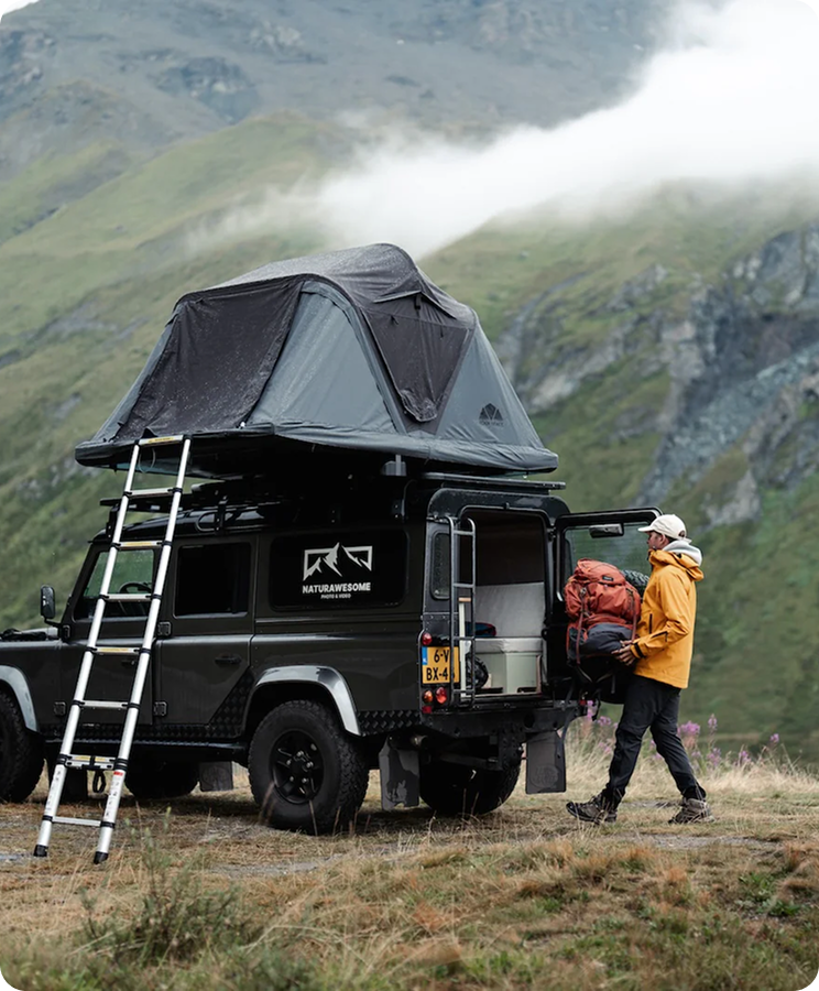 Ein Mann mit gelber Jacke entlädt einen Rucksack aus einem schwarzen Land Rover mit Dachzelt vor nebliger Bergkulisse. Schriftzug "NATURAWESOME" sichtbar.